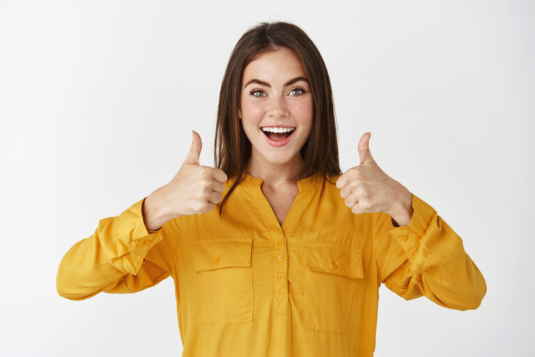 Impressed young woman showing thumbs up and smiling amazed, praising something cool, standing over white background